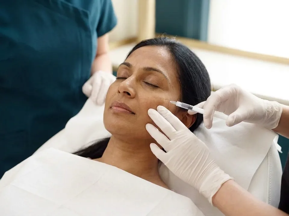 A relaxed woman lying back with her eyes closed while a white-gloved hand holds a small syringe with a very short, fine needle at her upper cheek