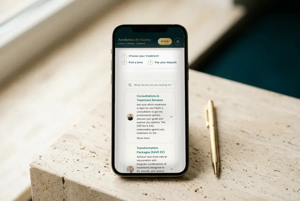 A phone standing on travertine showing the Aesthetics by Hayley booking page, beside a gold pen